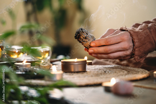 Woman hands burning white sage, before ritual on the table with candles and green plants. Smoke of smudging treats pain and stress, clear negative energy and meditation