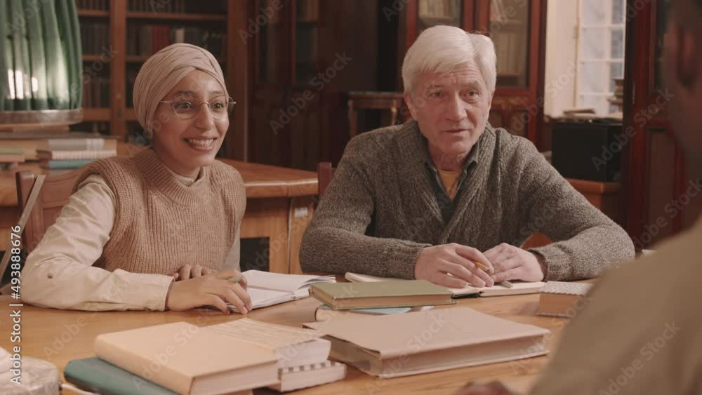 Waist up slowmo of diverse group of people discussing books sitting together at wooden desk in library Waist up slowmo of diverse group of people discussing books sitting together at wooden desk in library
