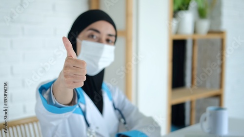 A medical worker shows a thumbs up in the office where he receives patients. A Muslim woman works as a doctor. A student at a medical university is doing an internship at a hospital. Masked doctor.