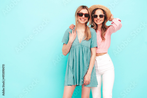 Two young beautiful smiling brunette hipster female in trendy summer dresses. Sexy carefree women posing near blue wall. Positive models having fun. Cheerful and happy. In hats and sunglasses