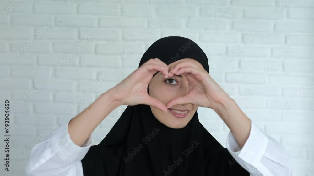 A Muslim woman in a black hijab smiles sweetly and shows a sign of love in the form of a heart folded with her fingers. A symbol of the Islamic world's love for the whole world. A Muslim woman in a black hijab smiles sweetly and shows a sign of love in the form of a heart folded with her fingers. A symbol of the Islamic world's love for the whole world.