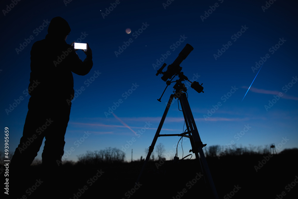 Silhouette of a man with smartphone, telescope and countryside under evening skies.