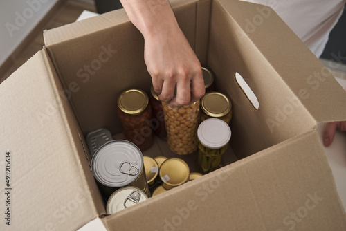 Unrecognizable young man filling donation box with food cans, legume jars and canned fish in kitchen.