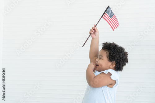 Adorable African American chubby kid girl smiling standing and holding small USA flags in hand at home. Celebrating 4th July Independence Day. Copy space. Side view. White background