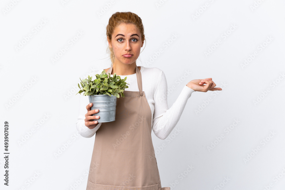 Young blonde gardener woman girl holding a plant over isolated white background pointing to the laterals having doubts Young blonde gardener woman girl holding a plant over isolated white background pointing to the laterals having doubts