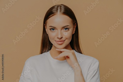 Lovely young European woman with makeup healthy skin touches gently jawline looks directly at camera has long straigh hair natural beauty dressed in casual sweater isolated over brown background photo