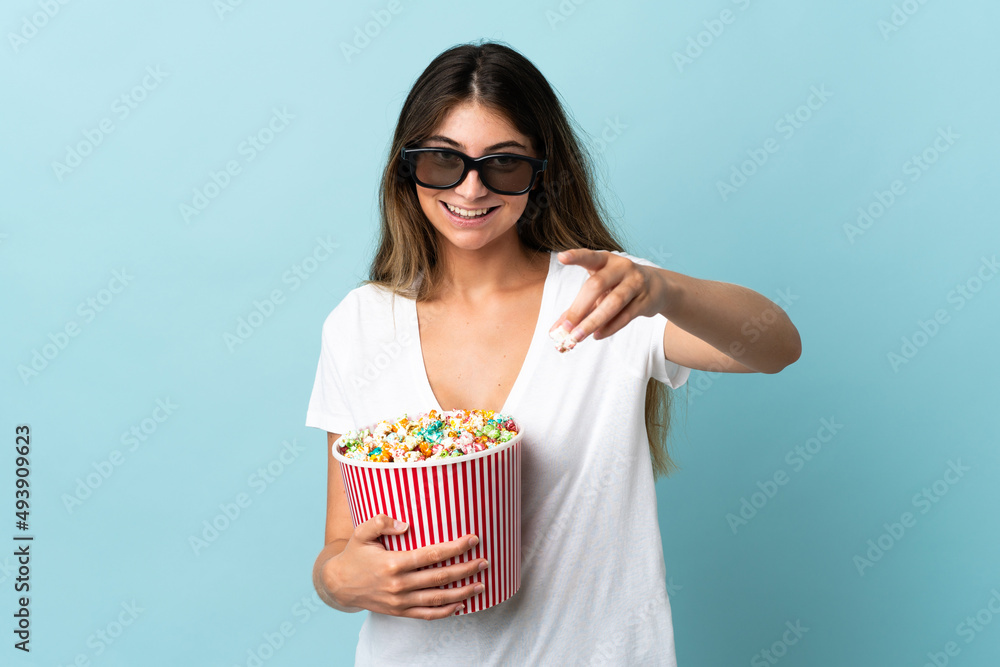 Young caucasian woman isolated on blue background with 3d glasses and holding a big bucket of popcorns while pointing front