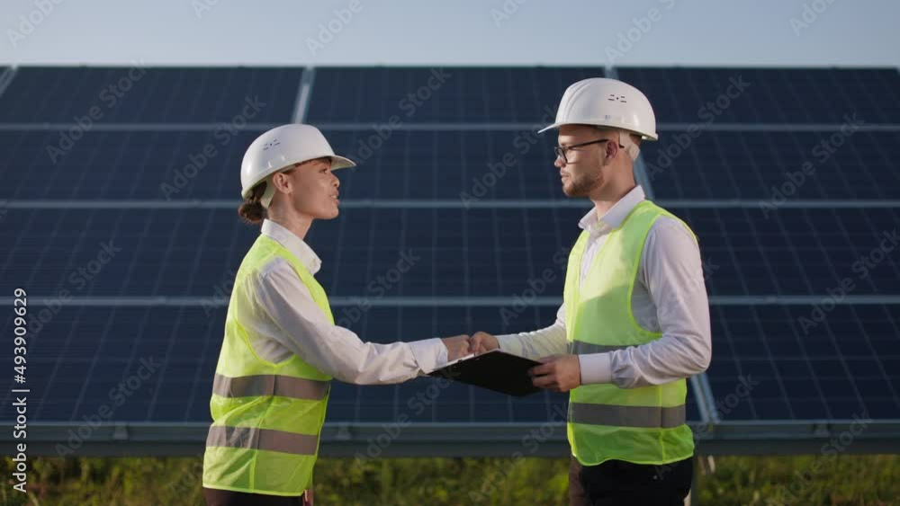 Side view of caucasian man and woman shaking hands for greeting each other during meeting on field with solar panels. Two coworkers in uniform doing inspection of modern station. Side view of caucasian man and woman shaking hands for greeting each other during meeting on field with solar panels. Two coworkers in uniform doing inspection of modern station.