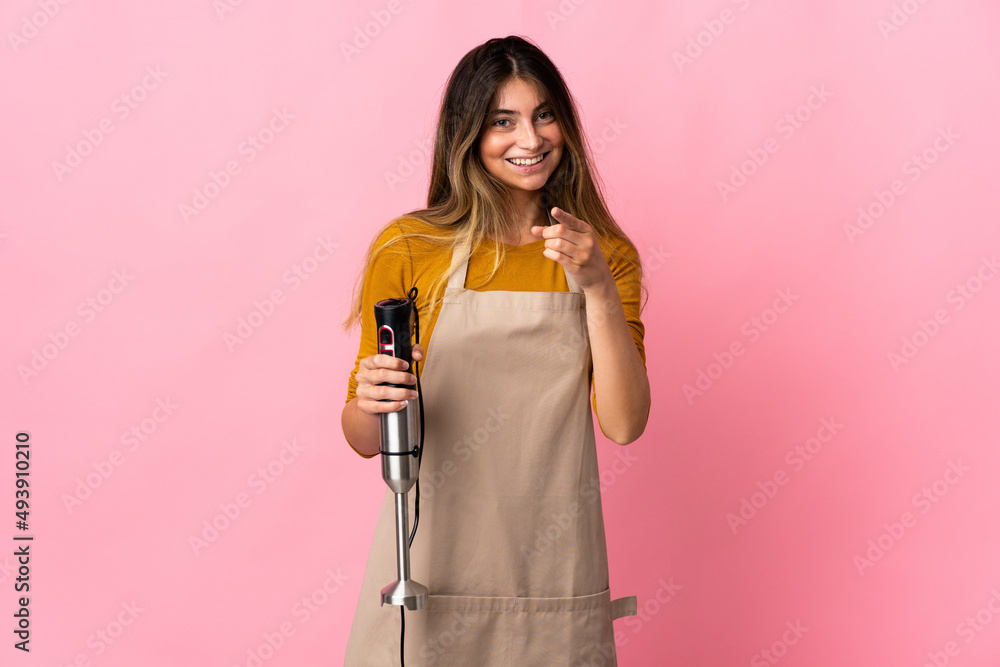 Young chef woman using hand blender isolated on pink background surprised and pointing front