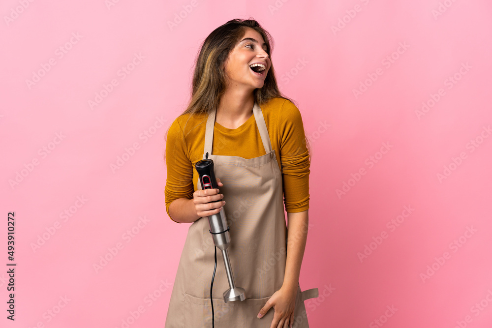 Young chef woman using hand blender isolated on pink background laughing