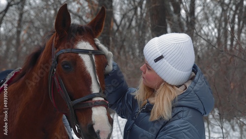 A young beautiful blonde in a hat, walking with a horse on a snowy winter field in the forest. A woman leads her horse by the bridle. She talks to animals and smiles. Horse breeding © stanis88