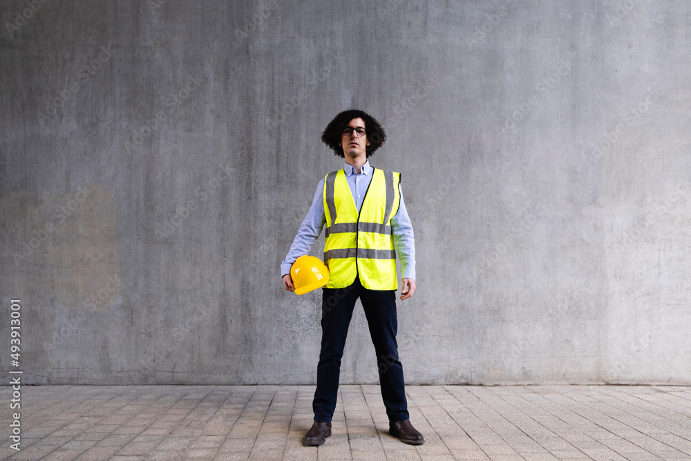 Man wearing reflective clothing holding hardhat standing in front of wall Man wearing reflective clothing holding hardhat standing in front of wall