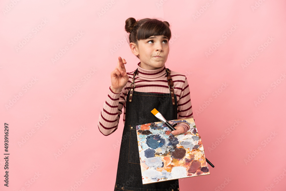 Little artist girl holding a palette isolated on pink background with fingers crossing and wishing the best Little artist girl holding a palette isolated on pink background with fingers crossing and wishing the best