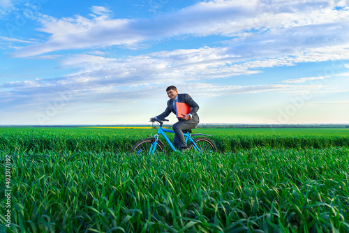 businessman with red folder for reports or documents rides a bicycle through a green grass field, dressed in a business suit, beautiful nature in spring, business concept