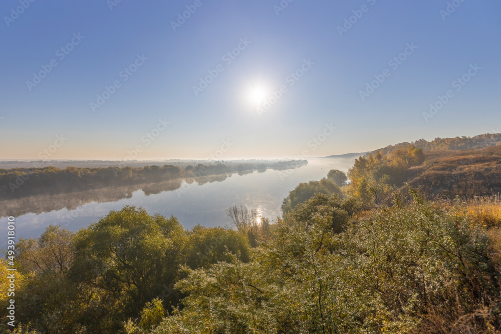 Autumn landscape in the early morning overlooking the river. A wide river and endless expanses of fields. Yellow leaves on trees and bushes are illuminated by the rays of the rising sun.