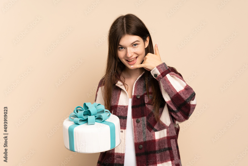 Young Ukrainian girl holding a big cake isolated on beige background making phone gesture. Call me back sign Young Ukrainian girl holding a big cake isolated on beige background making phone gesture. Call me back sign