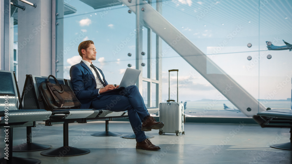 Airport Terminal: Businessman Uses Laptop Computer, Waiting for Flight. Traveling Man Relax after Work, Looking forward to Vacation. Sitting in a Boarding Lounge of Airline Hub with Airplanes Airport Terminal: Businessman Uses Laptop Computer, Waiting for Flight. Traveling Man Relax after Work, Looking forward to Vacation. Sitting in a Boarding Lounge of Airline Hub with Airplanes