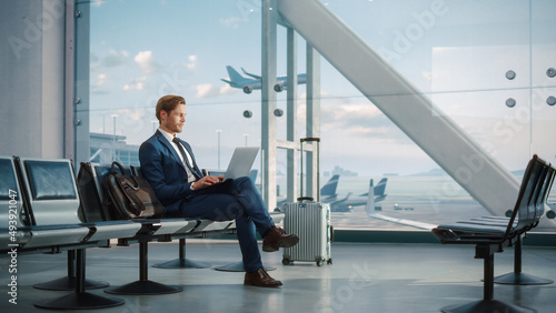 Modern Airport Terminal: Handsome Businessman Working on Laptop Computer While Waiting for His Flight. Man Sitting in a Boarding Lounge of Big Airline Hub with Airplanes Departing and Arriving photo