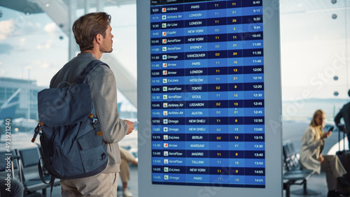 Airport Terminal: Young Man Looking at Arrival and Departure Information Display Looking for His Flight. Backgrond: Diverse Crowd of People Wait for their Flights in Boarding Lounge of Airline Hub