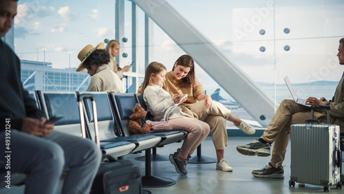 Busy Airport Airplane Terminal: Happy Beautiful Mother and Cute Little Daughter Wait for their Vacation Flight, Use Mobile Smartphone for Fun. Diverse Group of People in Boarding Lounge of Airline Hub