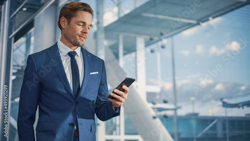 Airport Terminal: Businessman Walks to His Flight Gates, Uses Smartphone, Doing e-Business, Browsing the Internet with App. Digital Entrepreneur Remote Work Online While Traveling Through Airline Hub