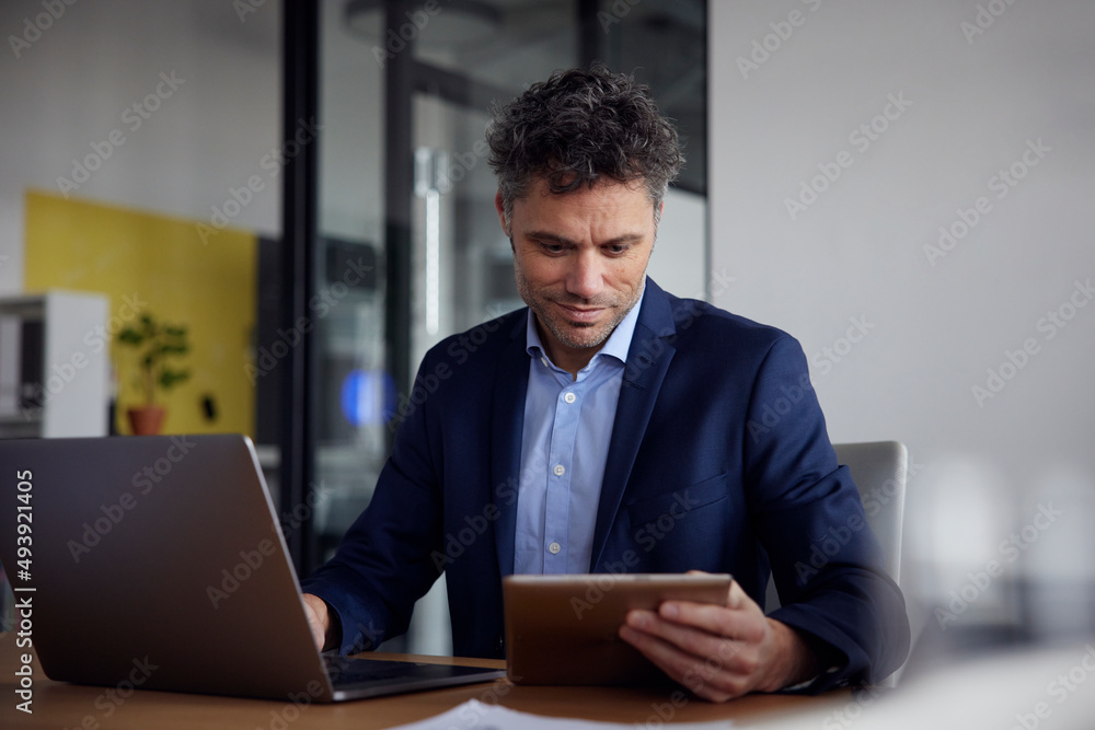 Smiling businessman using tablet PC sitting with laptop at desk in office Smiling businessman using tablet PC sitting with laptop at desk in office