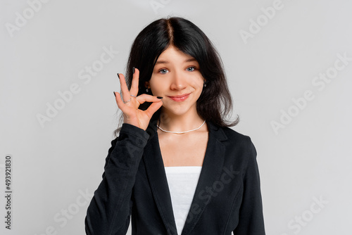 Your secrets safe with me concept. Close up portrait of young dark-haired girl showing zip gesture as if shutting mouth on key, standing in black blazer over grey background. Keep a secret