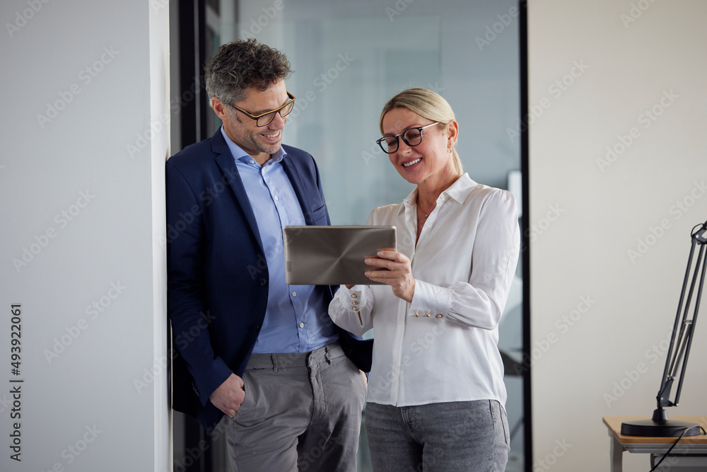 Colleague explaining over tablet PC to businessman leaning on wall with hand in pocket at office Colleague explaining over tablet PC to businessman leaning on wall with hand in pocket at office