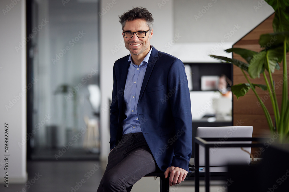 Smiling businessman wearing eyeglasses sitting on desk at work place Smiling businessman wearing eyeglasses sitting on desk at work place