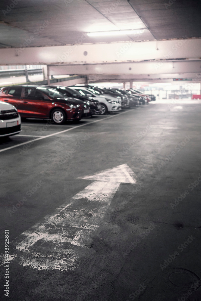 Directional white arrow on a floor of a parking lot and cars out of focus in the background. Selective focus. Busy car park of a shopping mall. Parking cost and vehicle density problem in town. Directional white arrow on a floor of a parking lot and cars out of focus in the background. Selective focus. Busy car park of a shopping mall. Parking cost and vehicle density problem in town.