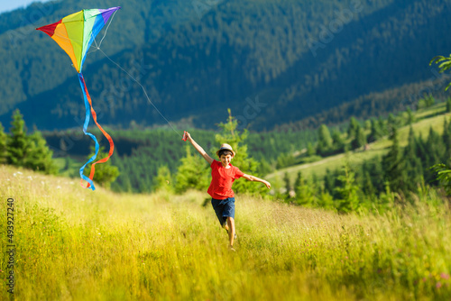 Happy child boy with a kite running on meadow in summer in mountains. Mountain landscape in the background with fir trees.