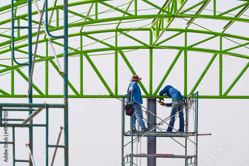 Asian foreman and construction worker on scaffolding are working to build metal roof structure of industrial building on lacking color sky background 