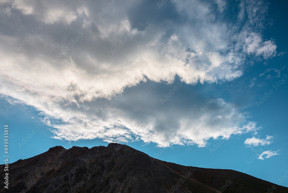 Scenic alpine landscape with dark mountain range silhouette under blue sky with spindrift clouds. Beautiful scenery with sunlit cirrus clouds above silhouette of mountain top at changeable weather.