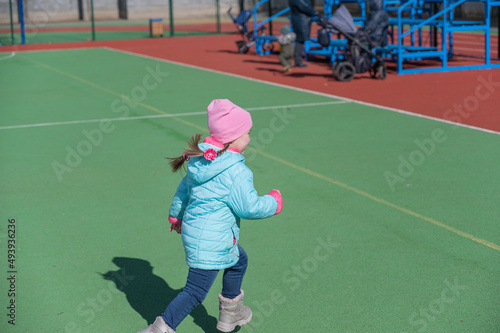 A child in a blue jacket runs around a green sports field. Parents and grandmother with baby carriages in the background. A mischievous five-year-old girl with pigtails frolicking. Childhood