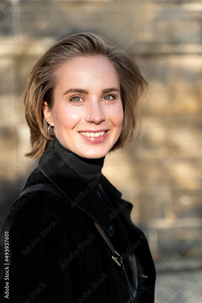 Portrait of young cheerful smiling blond short-haired woman with nose piercing, earrings, make up in velvet coat and black sweater having date walking outside on sunny day looking in camera. Weekend Portrait of young cheerful smiling blond short-haired woman with nose piercing, earrings, make up in velvet coat and black sweater having date walking outside on sunny day looking in camera. Weekend