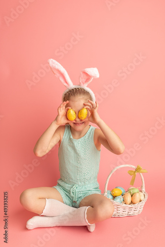 Portrait of cute smiling girl with Bunny ears and Easter basket, closes eyes with testicles, isolated on pink background. Happy Easter. Vertical photo