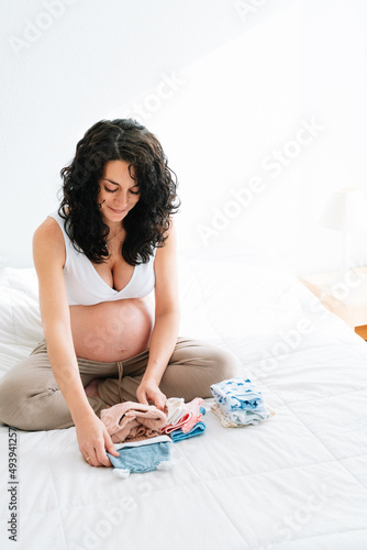 pregnant young woman sitting on bed folding and organizing clothes for her future baby