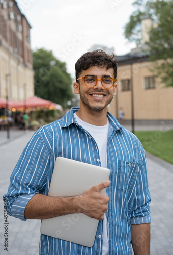 Authentic portrait of young handsome Indian man wearing stylish eyeglasses standing on the street. Smiling asian student holding laptop looking at camera in university campus. Education concept 