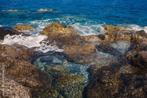 Wonderful views of the blue Mediterranean Sea. Sunny rocks, waves with foam and splashing water. The wave crashes into the rocks on the shore