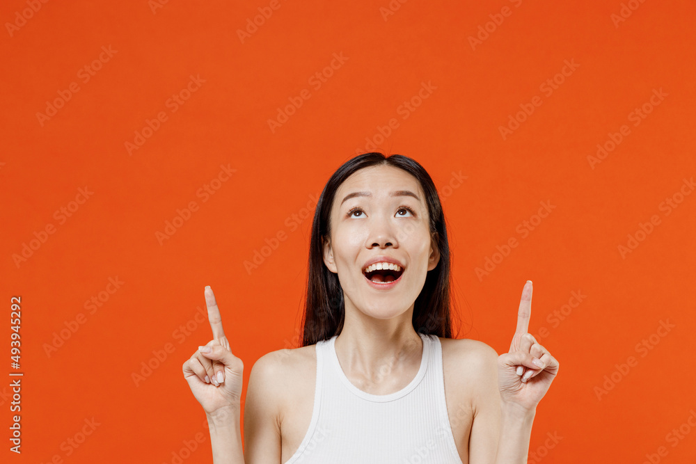 Overjoyed excited young woman of Asian ethnicity 20s years old in white tank top pointing forefingers overhead on workspace area copy space mock up isolated on plain orange background studio portrait. Overjoyed excited young woman of Asian ethnicity 20s years old in white tank top pointing forefingers overhead on workspace area copy space mock up isolated on plain orange background studio portrait.
