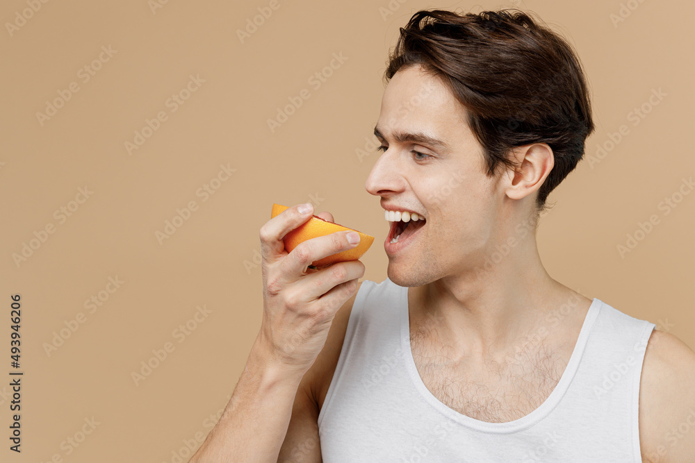 Attractive young man 20s perfect skin in undershirt hold in hand bite half of grapefruit isolated on pastel pastel beige background studio portrait. Skin care healthcare cosmetic procedures concept.