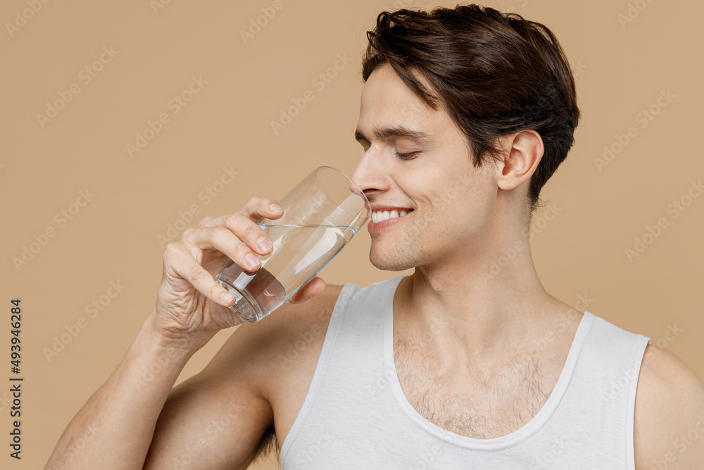 Attractive caucasian young man 20s perfect skin wear undershirt hold glass of water drink isolated on pastel pastel beige background studio portrait. Skin care healthcare cosmetic procedures concept. Attractive caucasian young man 20s perfect skin wear undershirt hold glass of water drink isolated on pastel pastel beige background studio portrait. Skin care healthcare cosmetic procedures concept.