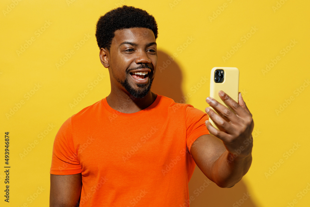 Young fun cool man of African American ethnicity 20s wearing orange t-shirt doing selfie shot on mobile cell phone post photo on social network isolated on plain yellow background studio portrait Young fun cool man of African American ethnicity 20s wearing orange t-shirt doing selfie shot on mobile cell phone post photo on social network isolated on plain yellow background studio portrait