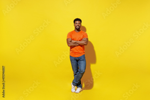 Full body young smiling fun happy man of African American ethnicity 20s in basic orange t-shirt hold hands crossed folded isolated on plain yellow background studio portrait. People lifestyle concept.