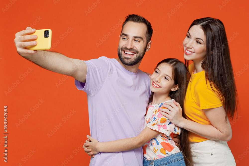 Young parents mom dad with child kid daughter teen girl in basic t-shirts doing selfie shot on mobile cell phone isolated on yellow background studio portrait. Family day parenthood childhood concept