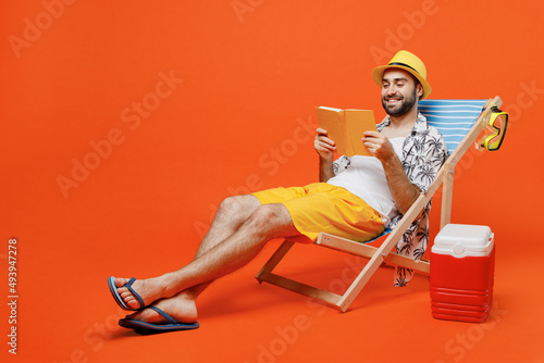 Photo Young smiling fun happy cool tourist man in beach shirt hat lie on deckchair near fridge read book novel isolated on plain orange background studio portrait