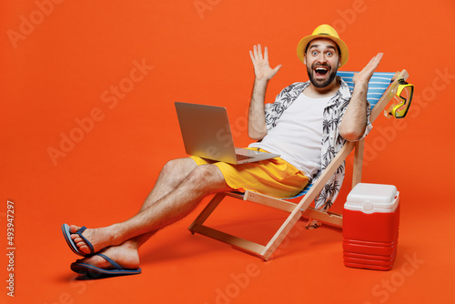 Fototapet Young surprised fun tourist man wear beach shirt hat lie on deckchair hold use work on laptop pc computer isolated on plain orange background studio portrait