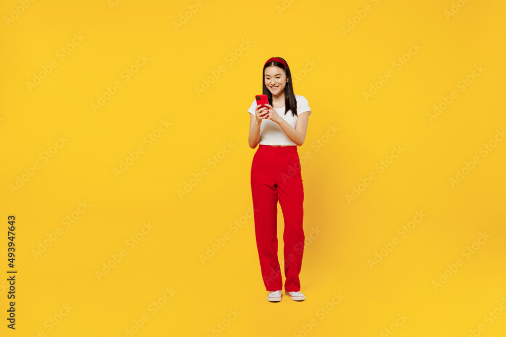 Full size body length smiling young girl woman of Asian ethnicity 20s years old wears casual clothes hold in hand use mobile cell phone send sms isolated on plain yellow background studio portrait Full size body length smiling young girl woman of Asian ethnicity 20s years old wears casual clothes hold in hand use mobile cell phone send sms isolated on plain yellow background studio portrait