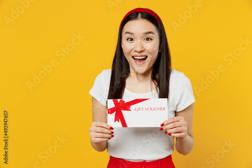 Cheerful smiling shocked young girl woman of Asian ethnicity 20s years old wears white t-shirt hold gift certificate coupon voucher card for store isolated on plain yellow background studio portrait.