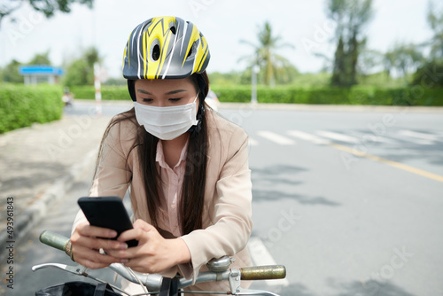 Young woman in protective helmet and medical mask sitting on bicycle and answering text messages on smarphone